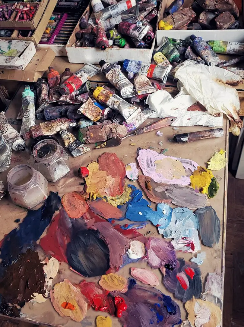 Vista superior de uma mesa de estúdio de artista desarrumada, com tubos de tinta a óleo usados, espátulas e uma paleta colorida coberta de tintas misturadas.