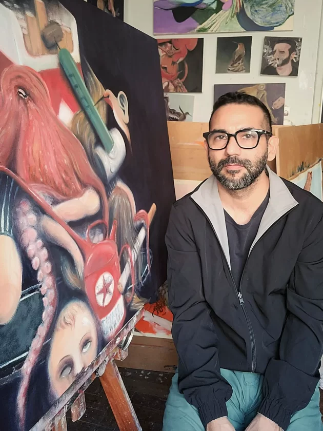 O artista Dario Velasco sentado em seu ateliê, ao lado de uma grande pintura surrealista de um polvo, com outras obras ao fundo.